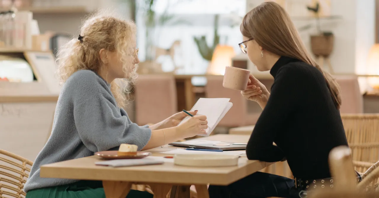 Two women discussing plans over coffee, reviewing notes and strategies in a cozy café setting.