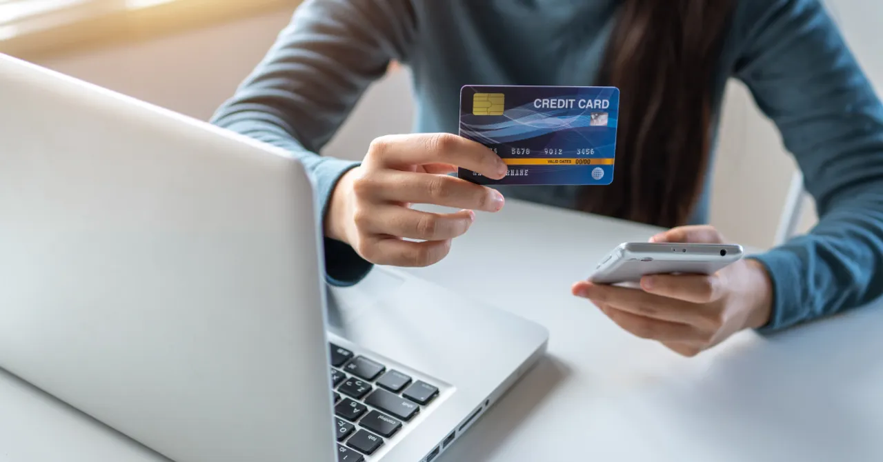 Person holding a credit card and smartphone while shopping online using a laptop.