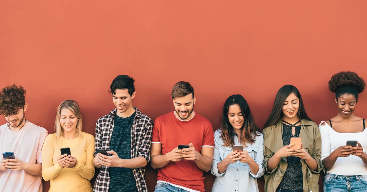 A group of happy young people standing against a bright wall, all focused on their phones—perfectly capturing the mobile-first shopping vibe for your Shopify app launch this summer.