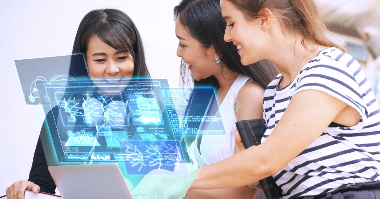 Three women smiling as they explore futuristic digital data displayed above a laptop, symbolizing technology-driven collaboration and innovation.