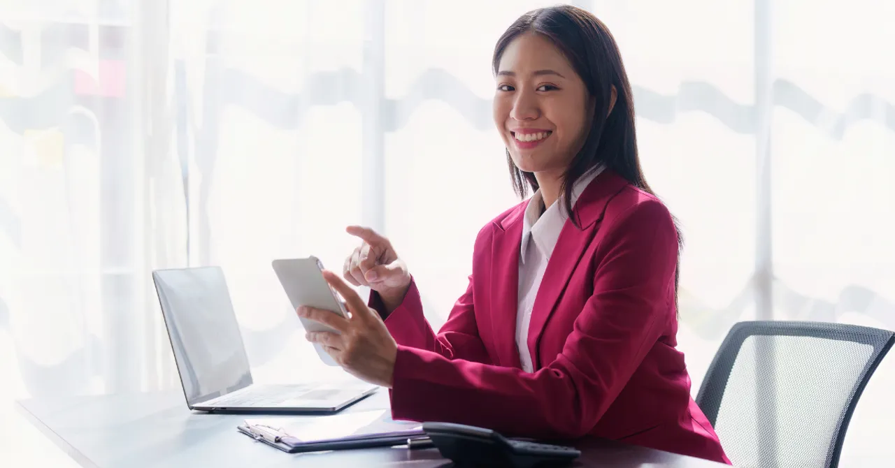 A confident businesswoman uses a smartphone and laptop at her desk, symbolizing how AI tools empower modern beauty brands on Shopify to connect with customers and drive sales smarter.