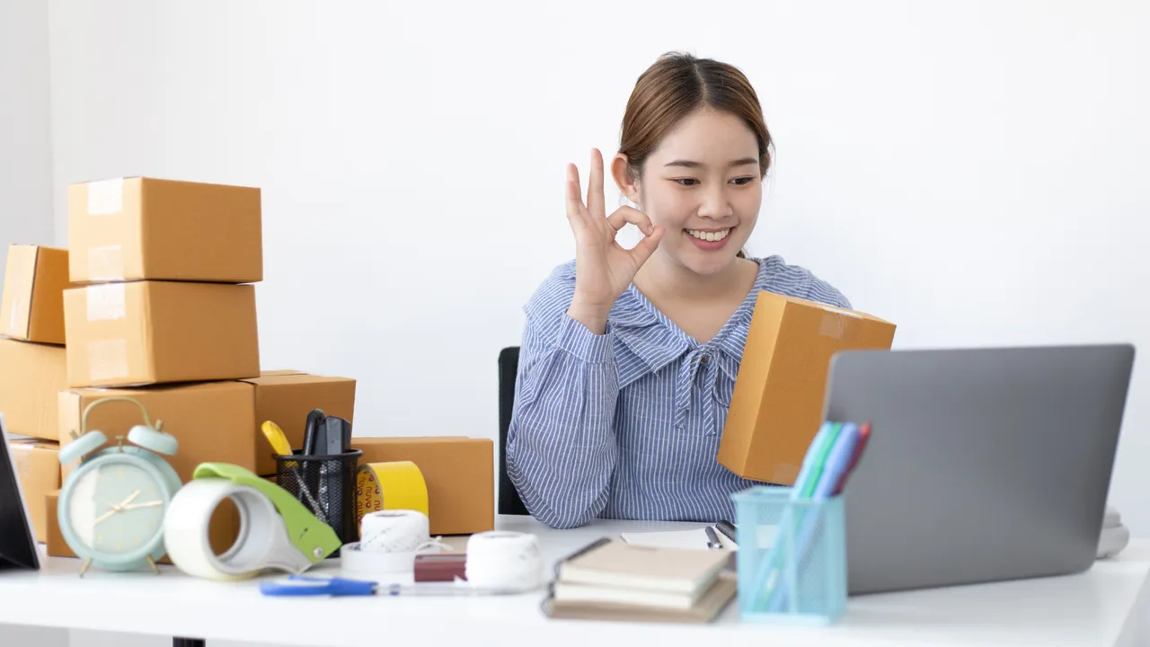 A smiling eCommerce seller holding a package and making an OK gesture in front of her laptop, surrounded by shipping boxes, representing the success and increased sales from using Supercart’s upsell solutions.  