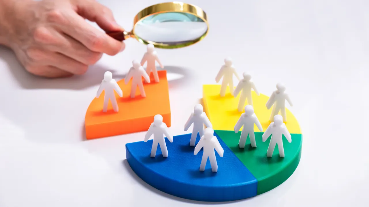 A hand holding a magnifying glass analyzing customer segments represented by colorful pie chart slices and miniature figures, symbolizing audience segmentation and targeted marketing.