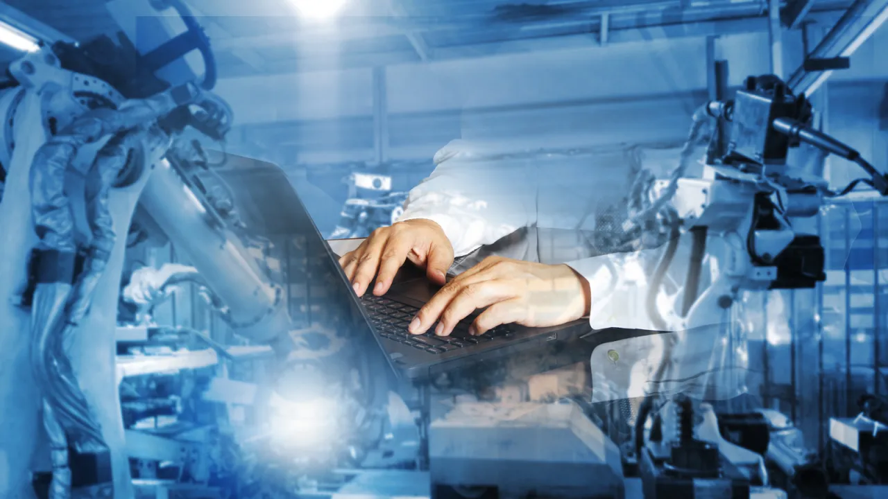 Hands typing on a laptop with a superimposed background of robotic arms in an industrial factory, symbolizing AI-driven automation and human-machine collaboration in modern manufacturing.