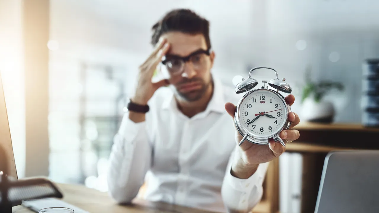 Frustrated businessman holding an alarm clock, symbolizing time pressure and poor time management at work.