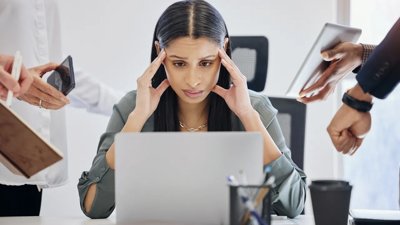 Stressed businesswoman surrounded by coworkers demanding attention while she works on her laptop.